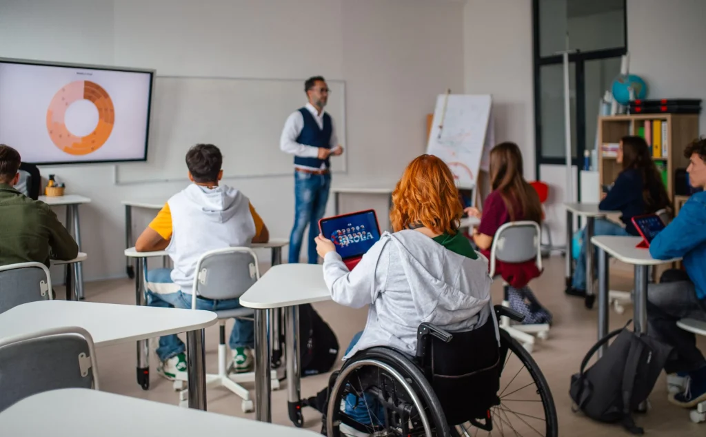 Students in a classroom, including a wheelchair user using a tablet, learning together and representing inclusive higher education.